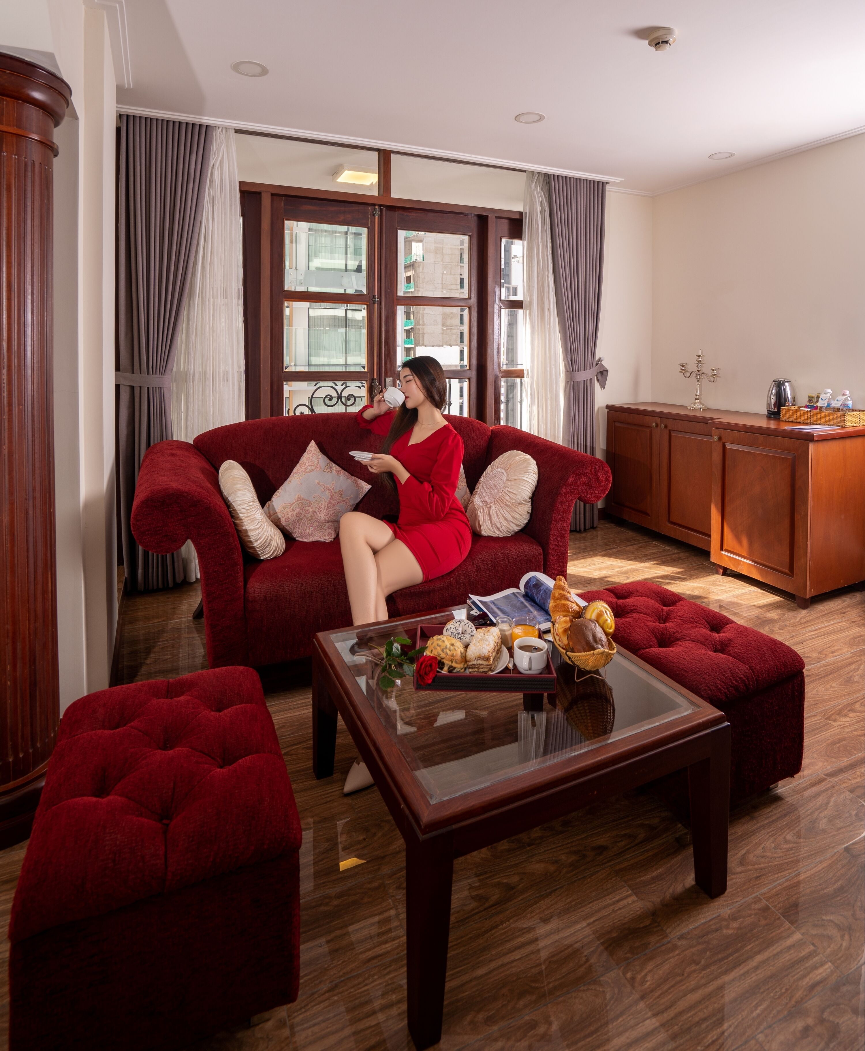 Una sala de estar con un sofá rojo, un reposapiés a juego, una mesa de centro de vidrio con alimentos para el desayuno y un armario de madera.