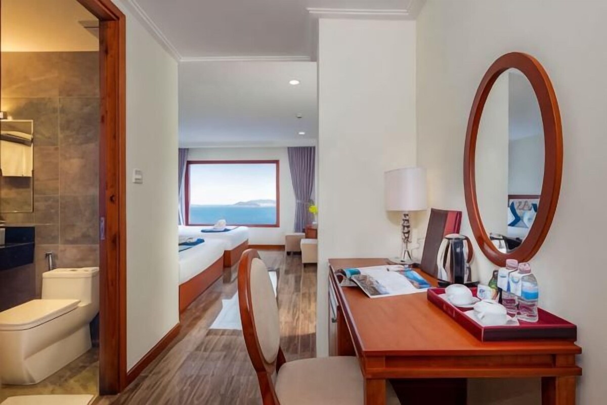 Habitación de hotel con cama, escritorio con espejo, silla, baño con inodoro y lavabo, y vista al mar.
