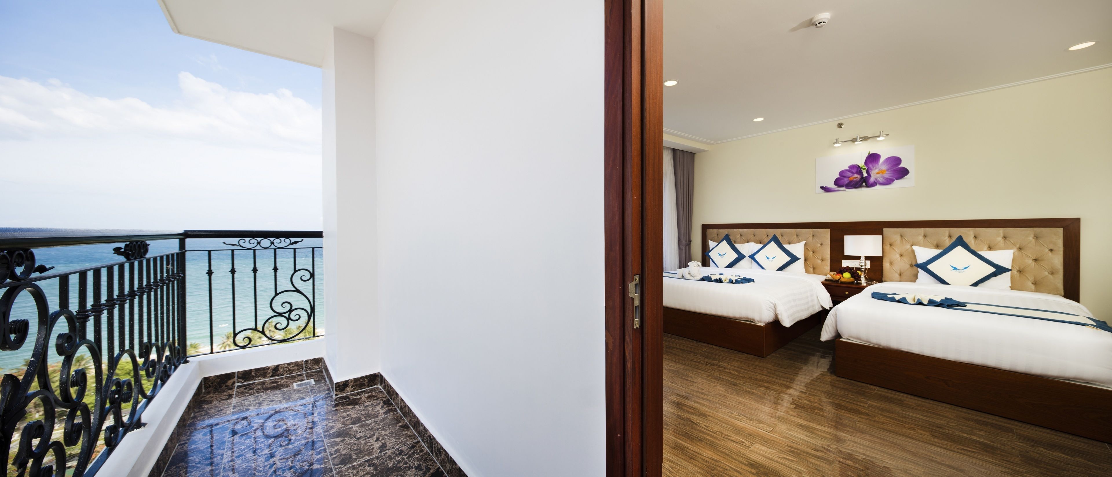 A hotel room with a balcony overlooking the sea, two beds with matching headboards, and a floral wall decoration.