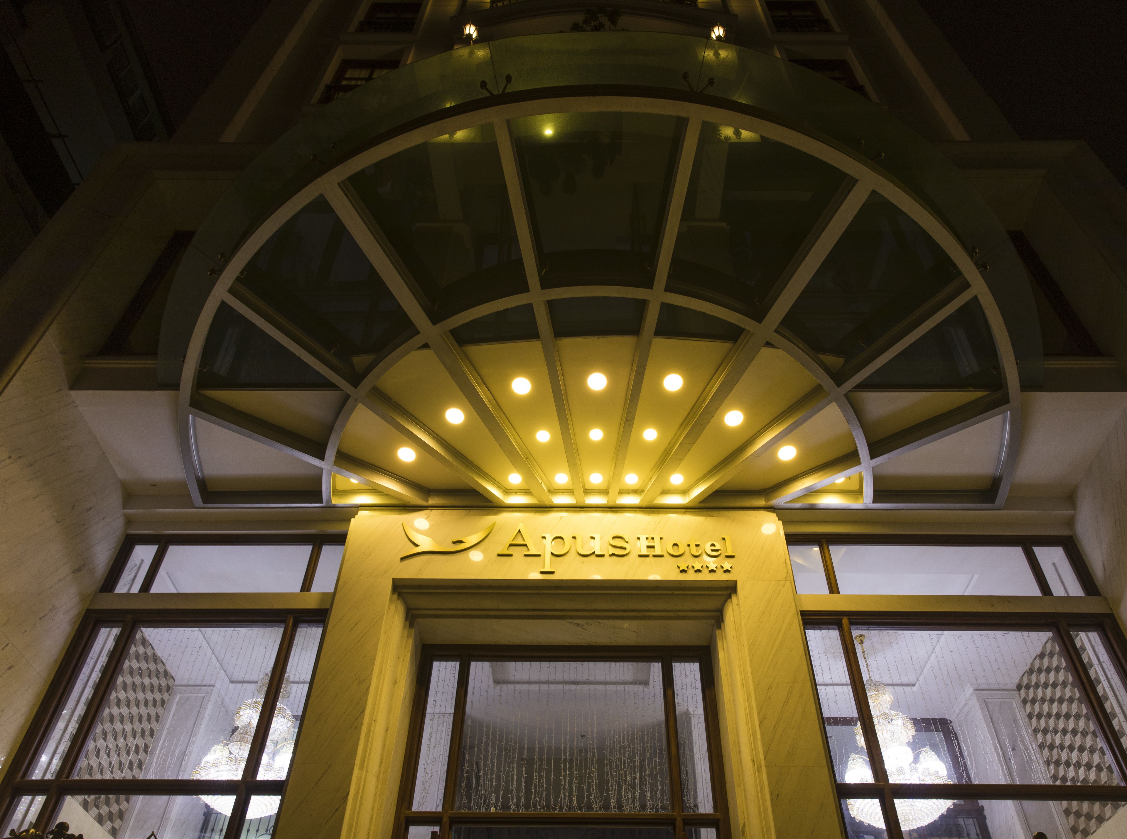 The entrance of the APUSH Hotel with a glass canopy and illuminated sign.