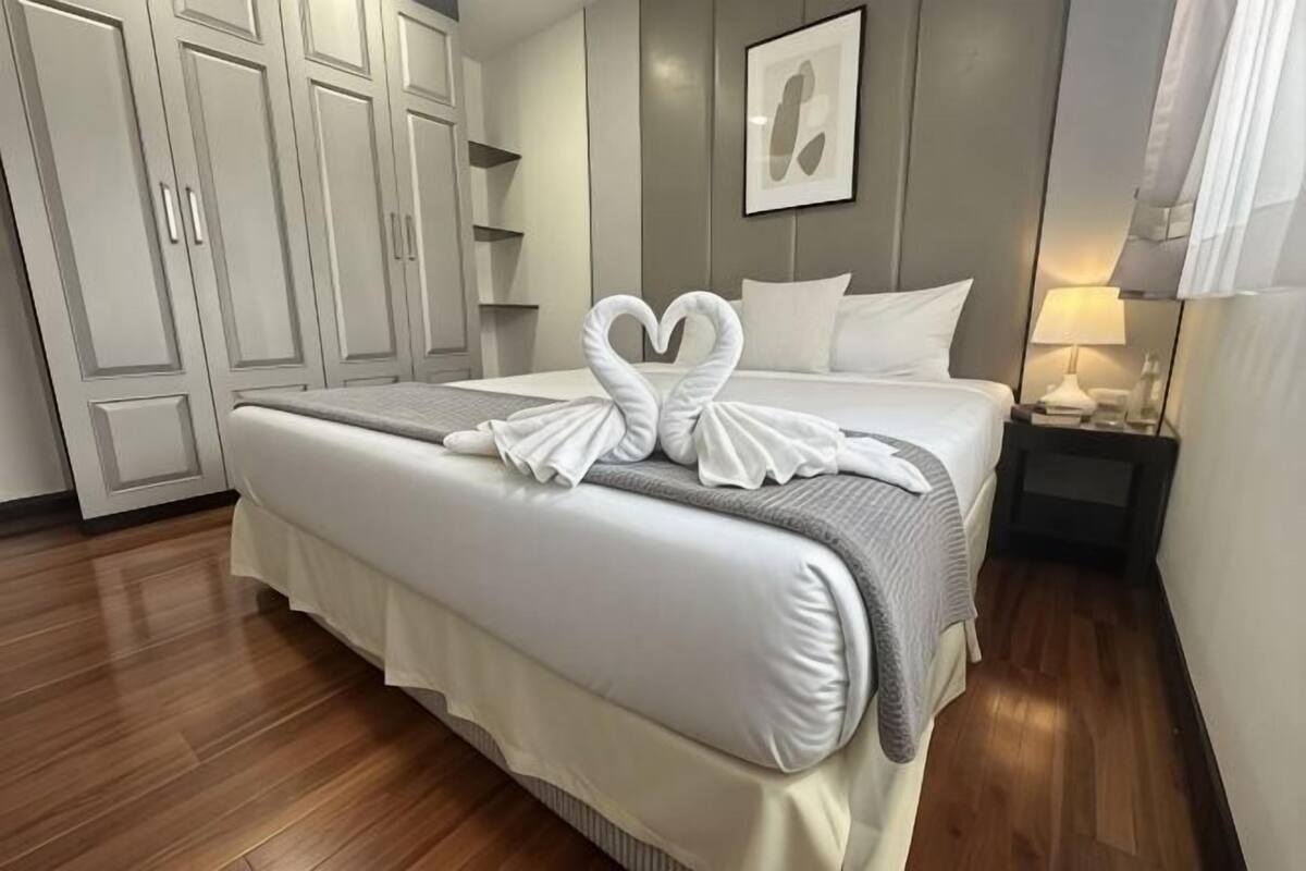 A neatly made bed with a heart-shaped towel arrangement, a bedside table with a lamp, and a wardrobe in a hotel room.
