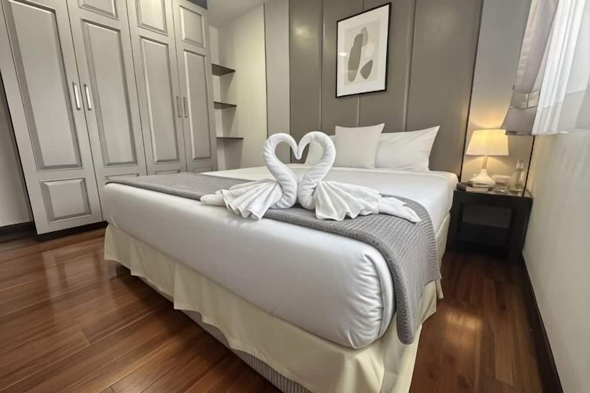 Un lit bien fait, agrĂ©mentĂ© dâune serviette pliĂ©e en forme de cĆur, une table de chevet avec une lampe et une armoire dans une chambre dâhĂŽtel.