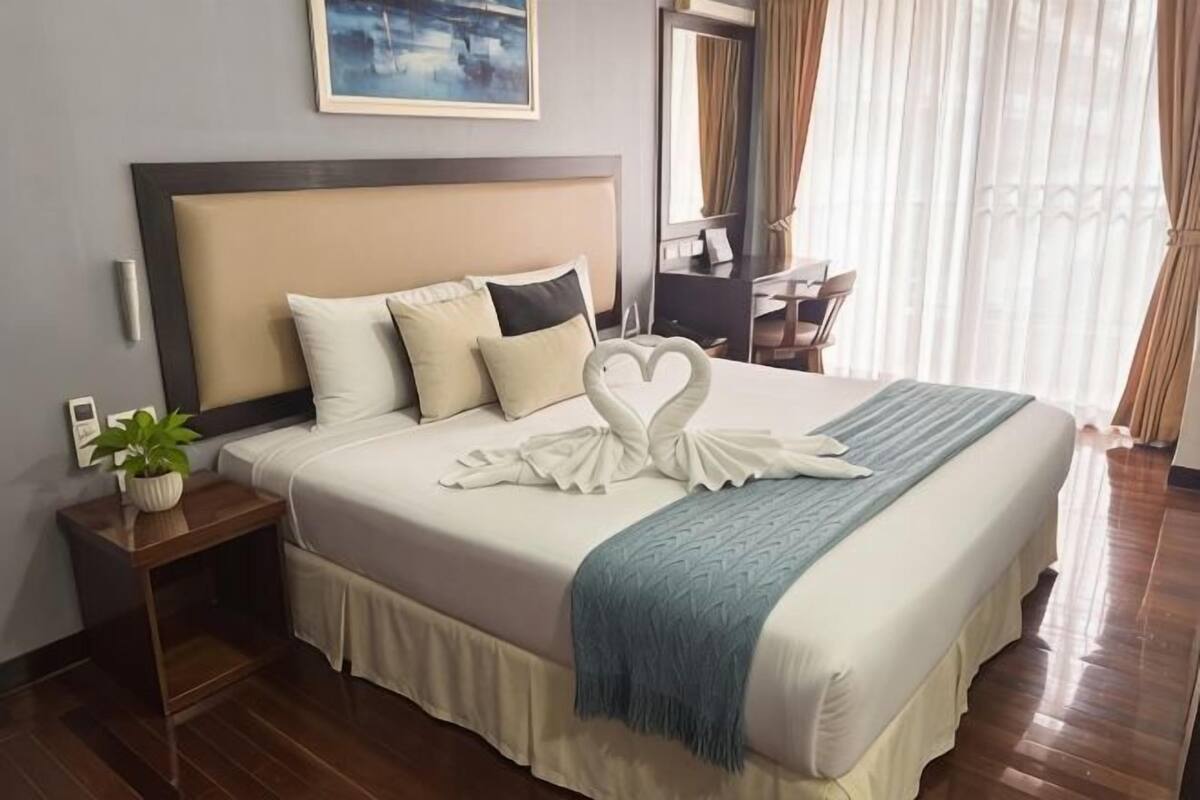 A neatly made bed with a heart-shaped towel, a bedside plant, and a blue throw blanket.