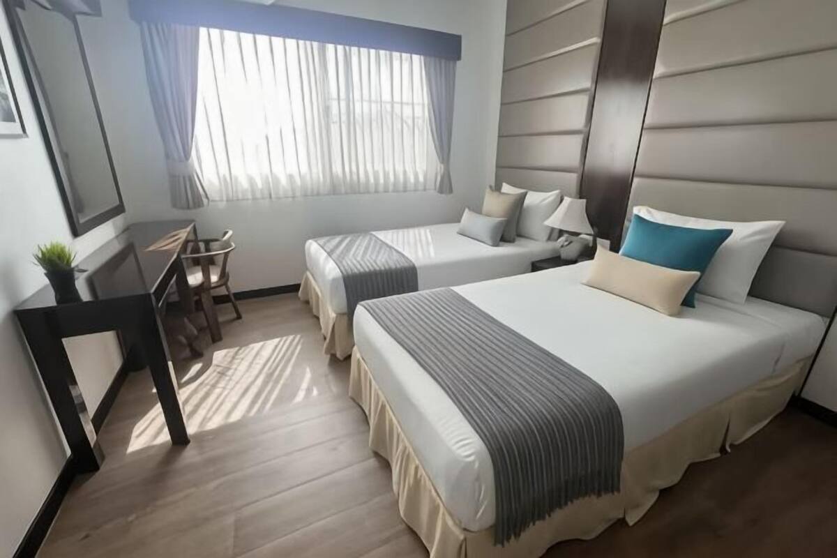 A hotel room with two beds, a desk, a chair, a window with curtains, and a mirror.