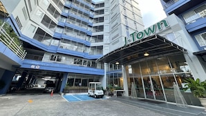 Un hotel de varios pisos con un llamativo letrero 'J-Town', una entrada cubierta y un estacionamiento con un vehículo blanco.