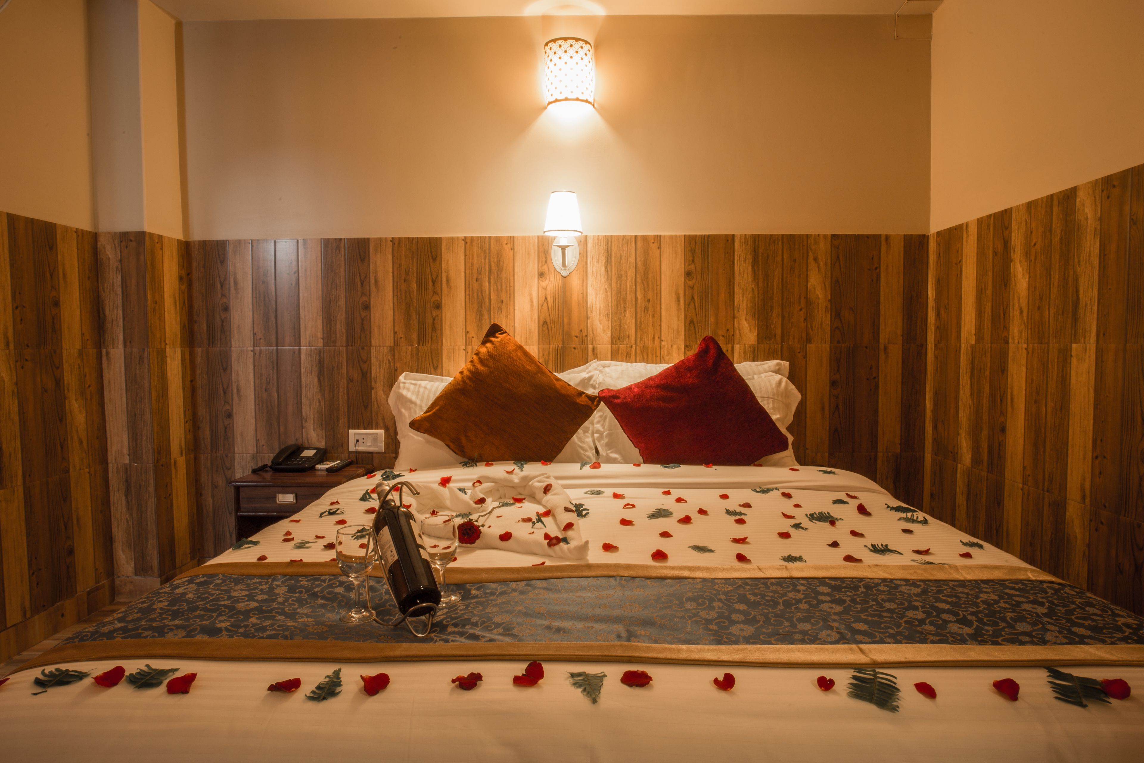 A hotel room with a wooden panel wall, a bed with a floral bedspread, a bedside lamp, and a nightstand with a phone and a glass.