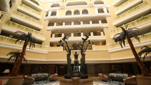 Um hall de hotel em vários níveis, com uma fonte central, palmeiras e um padrão de piso quadriculado.