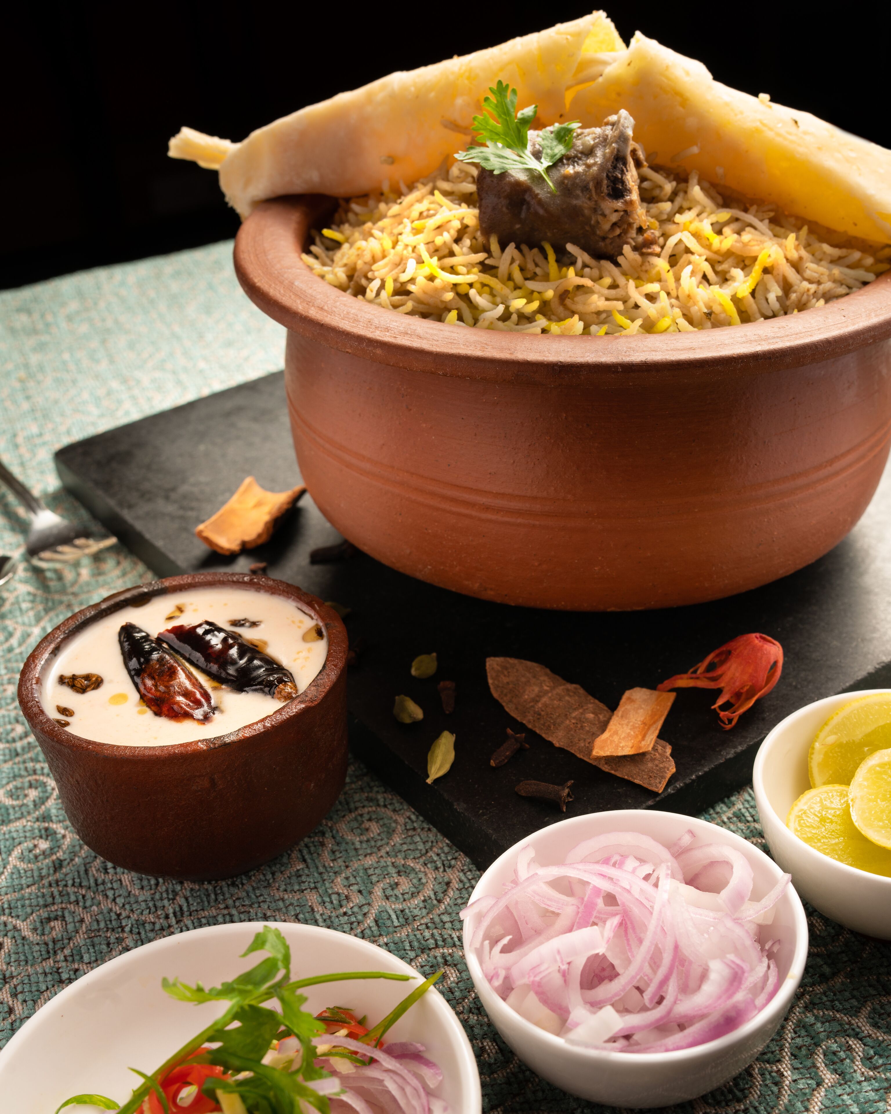 Ein traditionelles Biryani-Gericht, serviert in einem Terrakottatopf, begleitet von einem Joghurtbecher, geschnittenen Zwiebeln und Zitronenspalten.