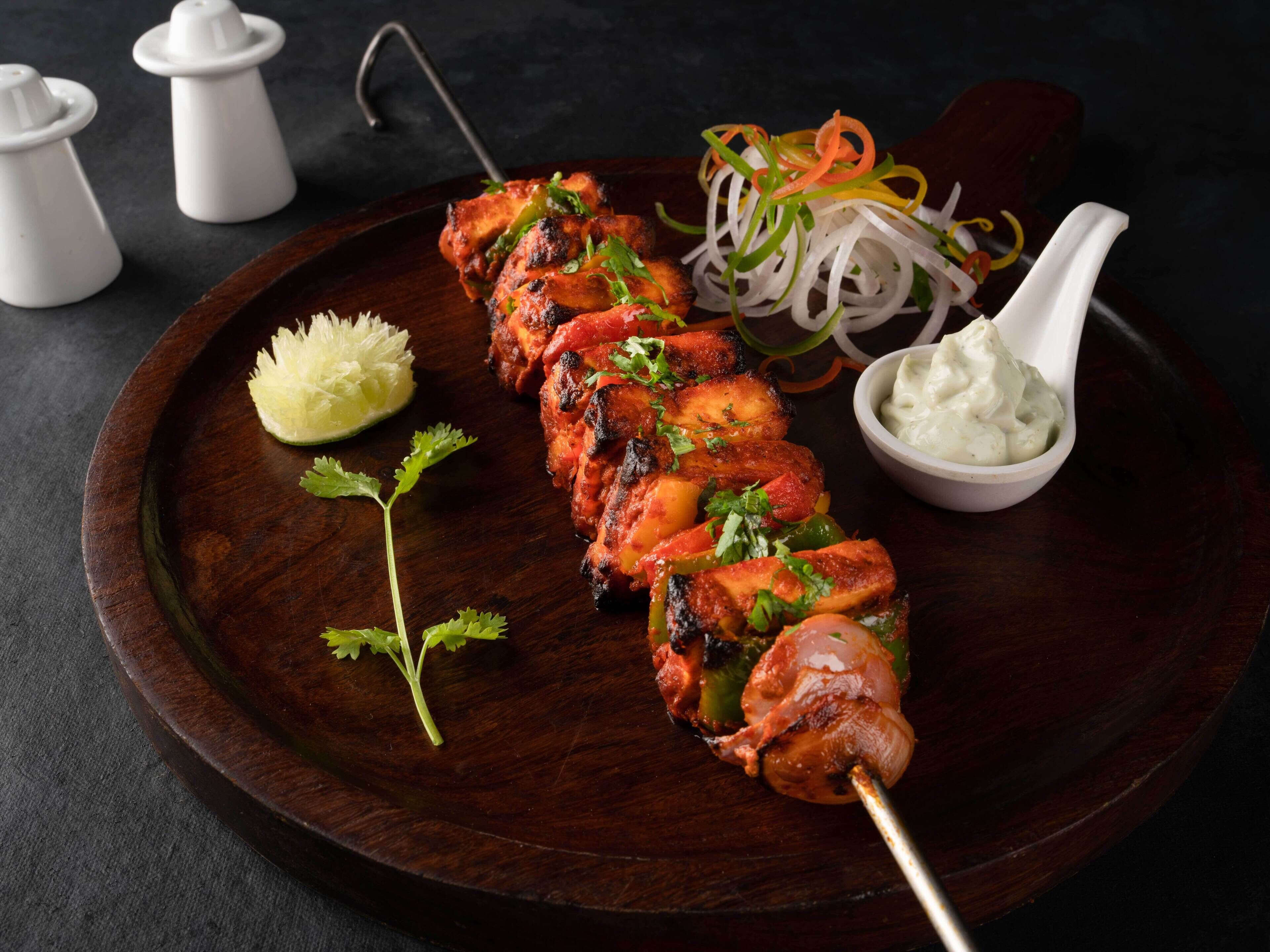 Un plato de madera con brochetas de pollo a la parrilla, un gajo de lima, y una guarnición de verduras en rodajas y salsa cremosa.