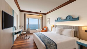 Quarto de hotel com uma cama grande, uma escrivaninha, uma cadeira, uma mesinha e vista para o mar.