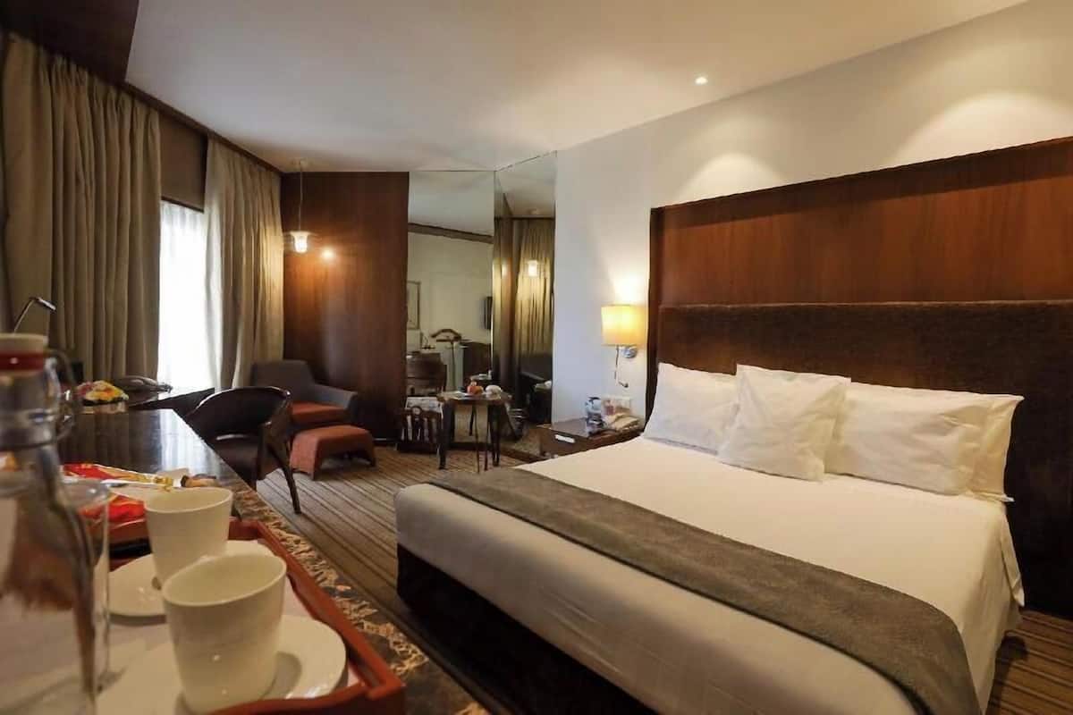 A hotel room with a bed, a dining table with a cup and saucer, a chair, a lamp, and a mirror.