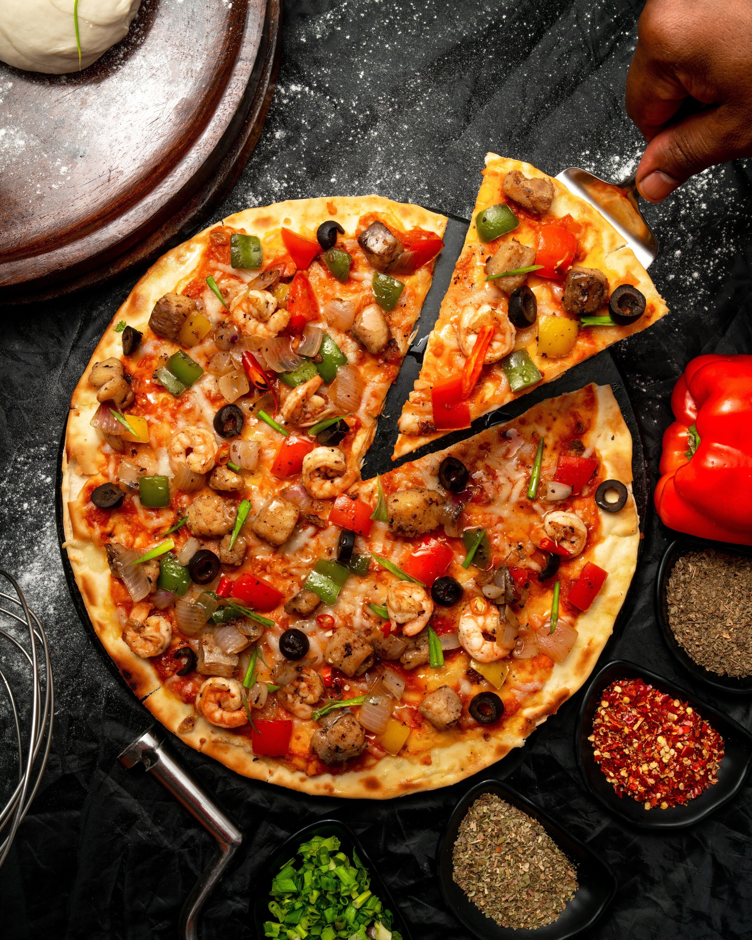 A pizza with various toppings, a pizza cutter, and some spices on a dark surface.