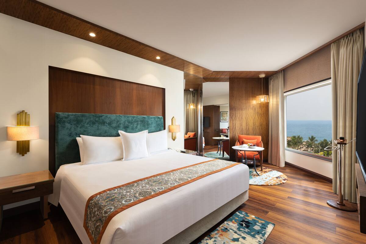 Habitación de hotel con una cama grande, vista al océano, un pequeño comedor y un baño con un espejo amplio.