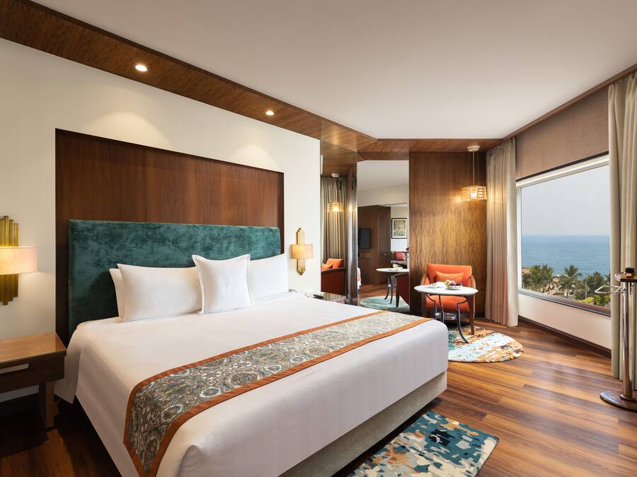 A hotel room with a large bed, a view of the ocean, a small dining area, and a bathroom with a large mirror.