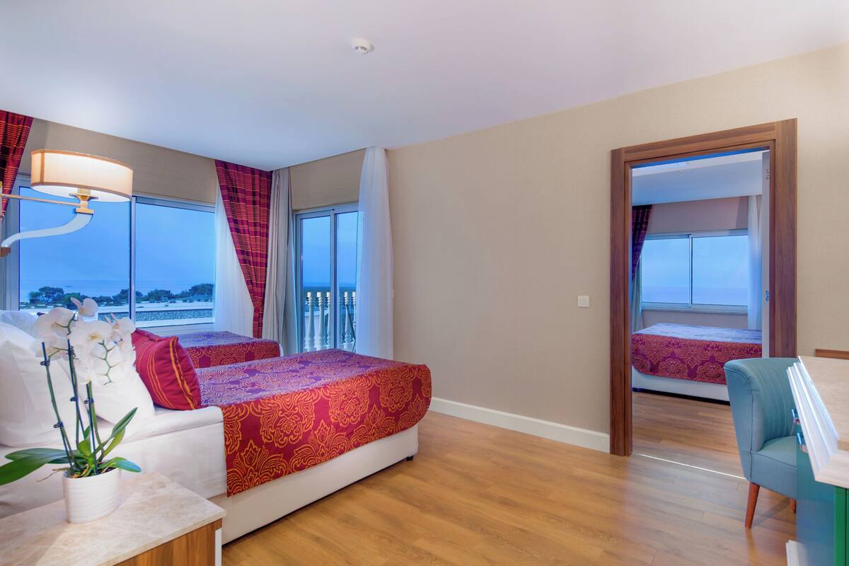 A hotel room with a bed, a desk, a chair, a potted plant, and a view of the outside.