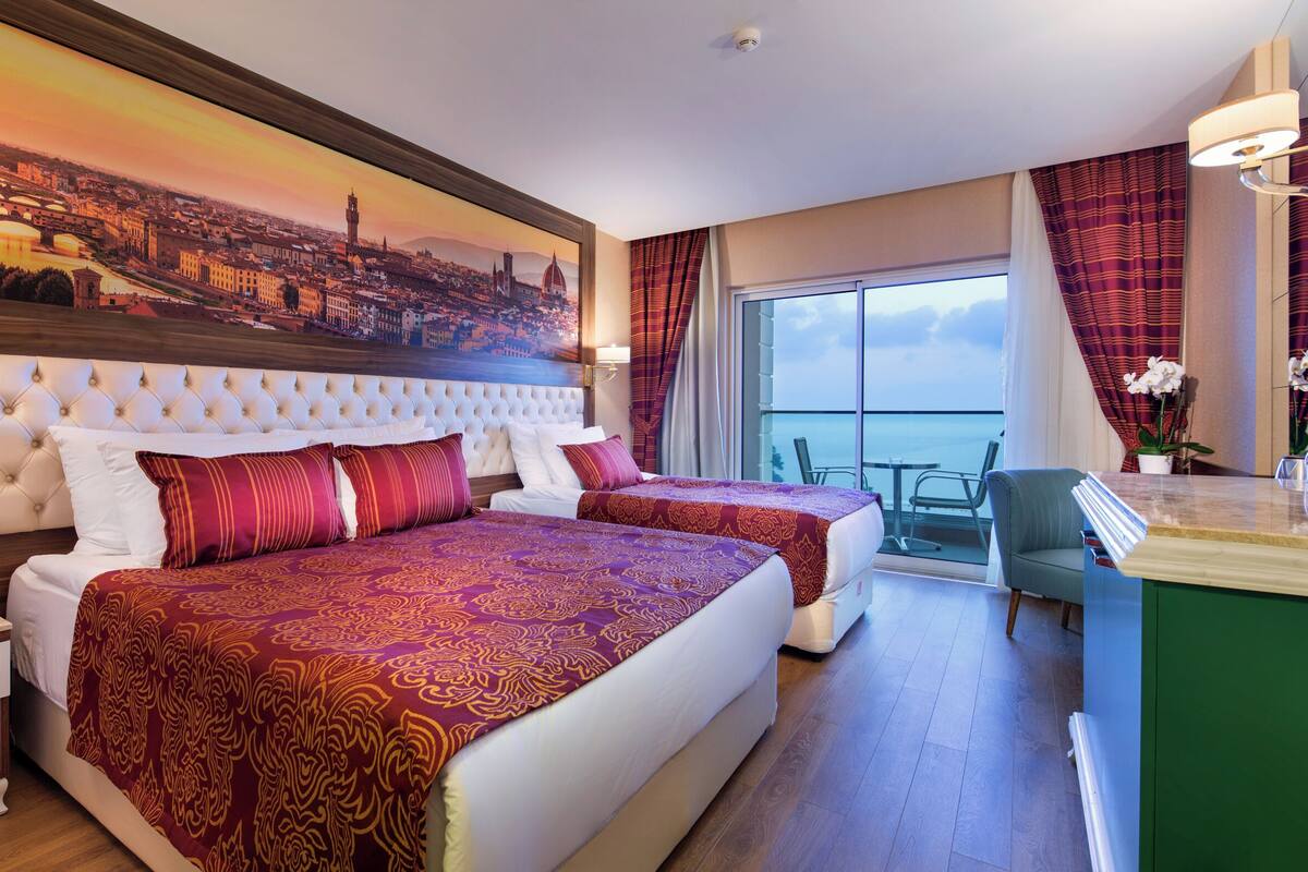 A hotel room with a large bed, a smaller bed, a balcony with a view, a desk, and a painting of a cityscape.