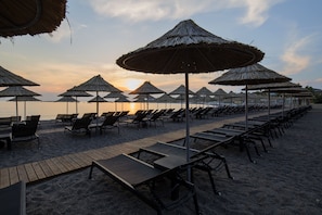 A beach with rows of sun loungers and thatched umbrellas, a wooden walkway, and a view of the sea.