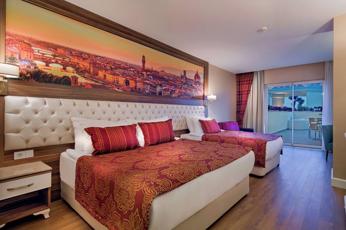 A hotel room with two beds, a large cityscape painting, a television, and a balcony with a view.