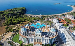 A large hotel with a central swimming pool, surrounded by lush greenery and a beach.