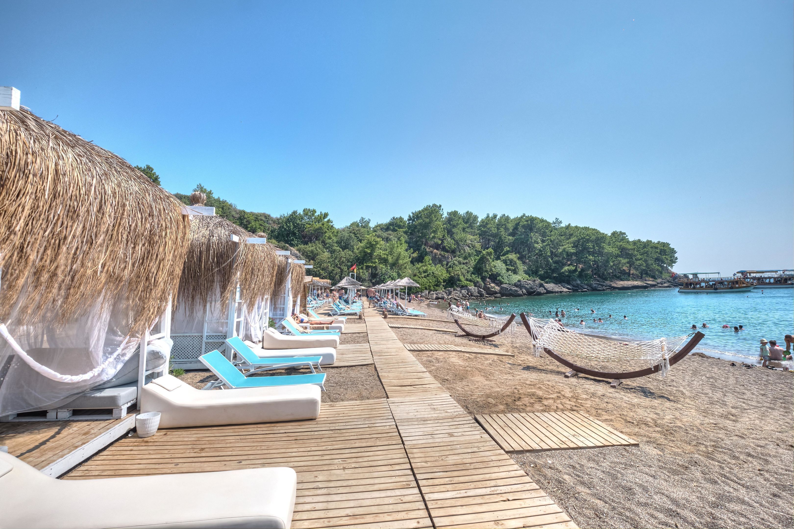 Una terraza de madera con sillones de descanso y una hamaca, una playa con aguas azules cristalinas y un promontorio boscoso al fondo.