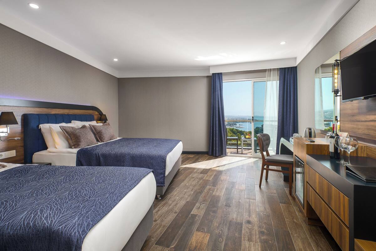 A hotel room with two beds, a desk, a chair, a TV, and a large window with a view.