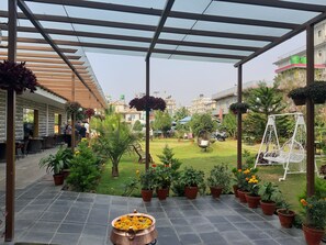 An outdoor patio with a covered seating area, potted plants, and a decorative copper pot with flowers. - Hotel Crown Himalayas (Pokhara)