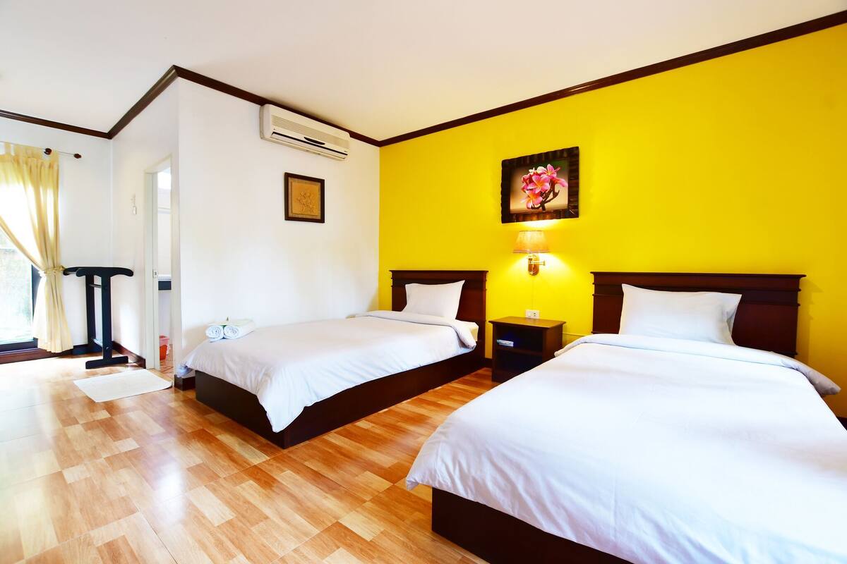 A hotel room with two beds, wooden flooring, a yellow accent wall, and a nightstand with a lamp.