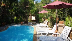A pool area with white lounge chairs and red umbrellas, surrounded by lush greenery. - Pathu Resort Ranong (Ranong)