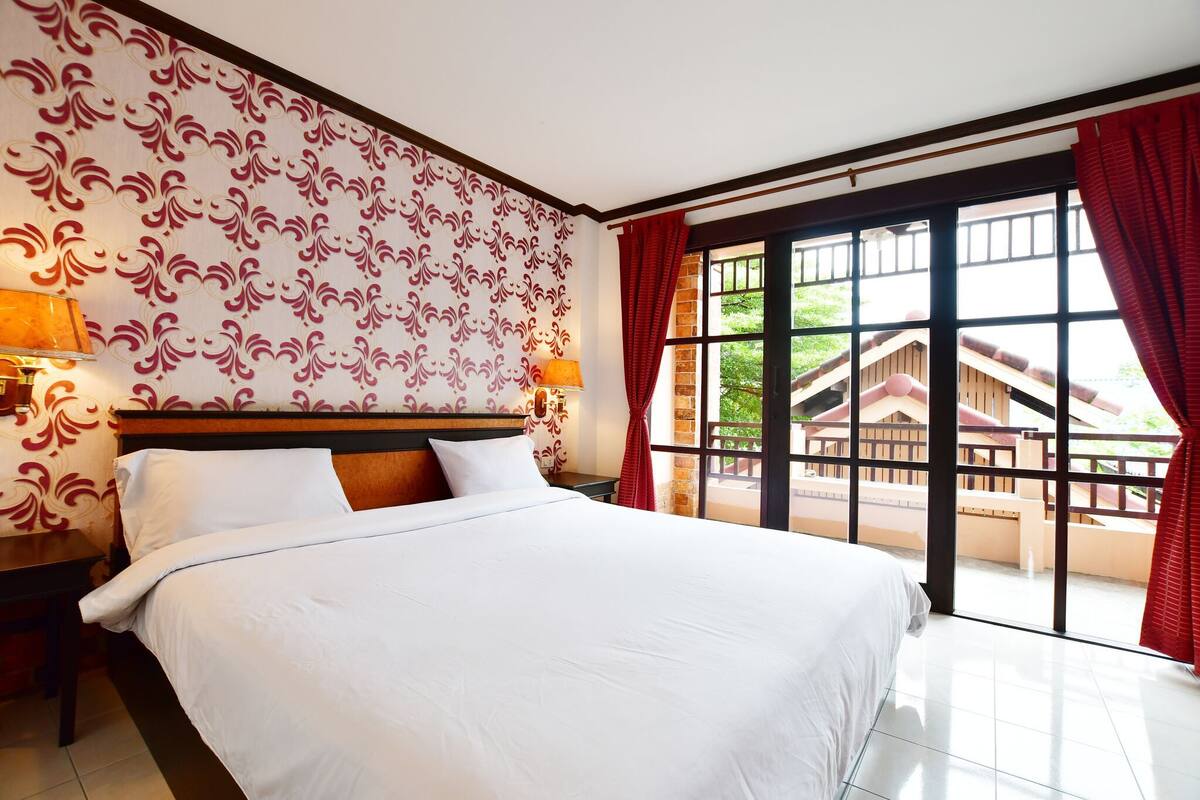 A hotel room with a large bed, a nightstand, a lamp, and a view of a wooden structure outside through a large window.