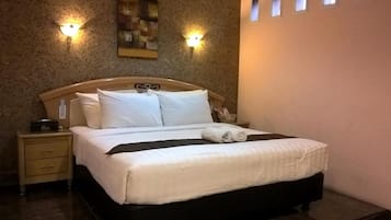 Una cama impecablemente hecha con sábanas blancas, un cojín negro decorativo y una toalla doblada a un lado. Hay dos mesitas de noche con lámparas, una de ellas con un objeto decorativo. La habitación tiene papel tapiz con motivos florales y una ventana con persianas.