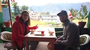 Twee personen zitten aan een houten tafel met kommen en een kopje, met uitzicht op een bergachtig landschap.