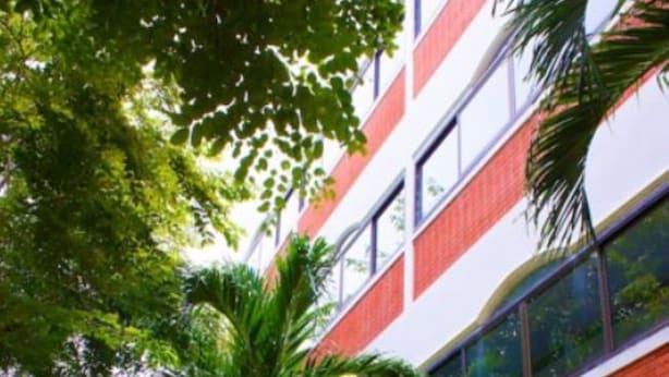 A multi-story building with a red and white facade, surrounded by lush greenery and palm trees.