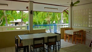 A covered outdoor dining area with a table and chairs, a blue fence, and a view of greenery.
