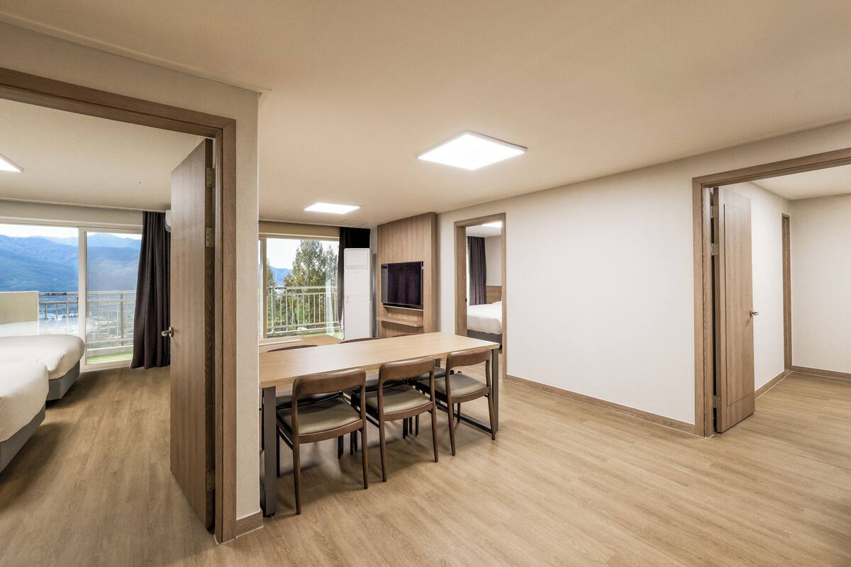Een hotelkamer met een houten vloer, een bed, een eettafel met stoelen, een televisie en een balkon met uitzicht op de bergen.