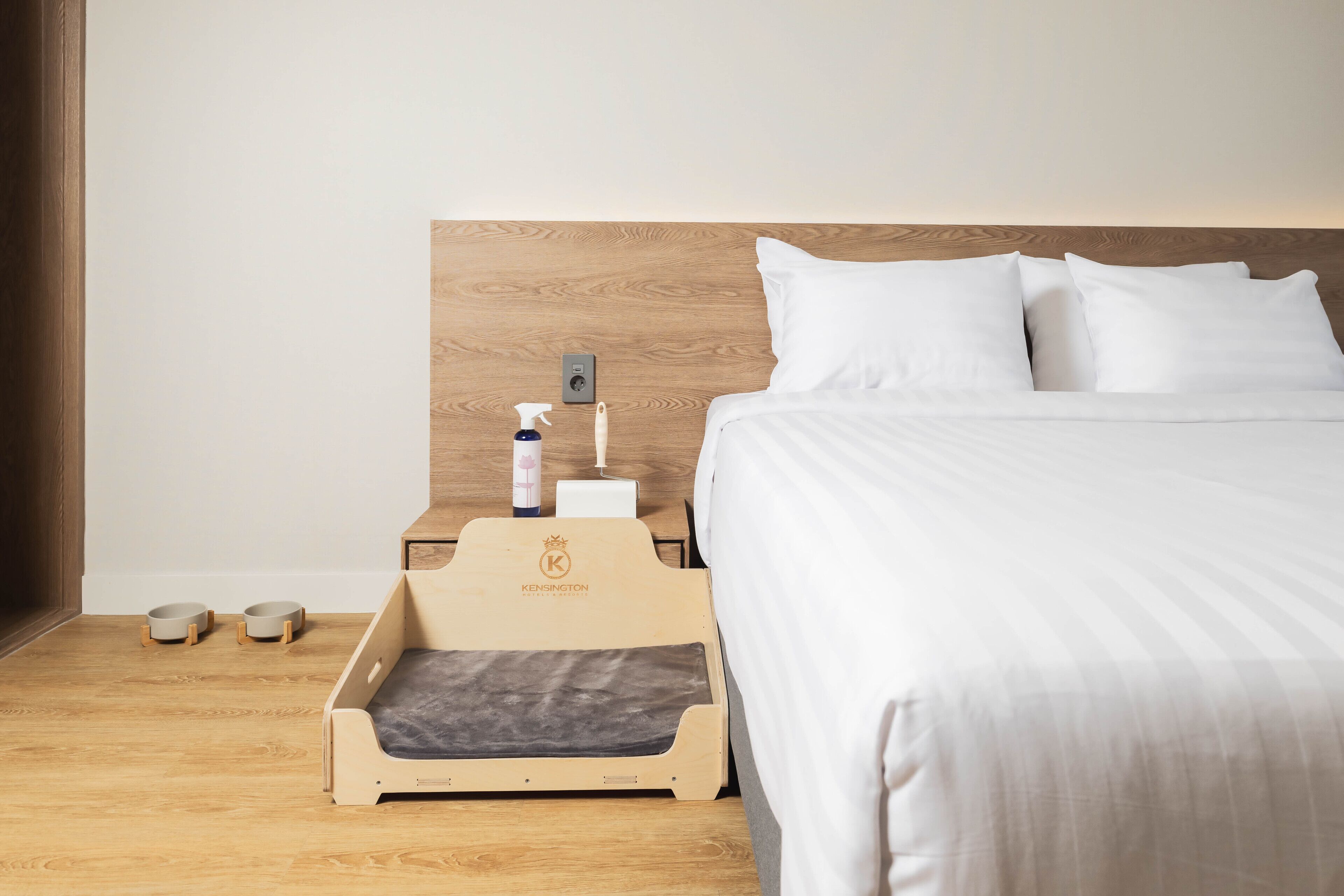 A hotel room with a wooden headboard, white bedding, a pet bed, and a small table with a spray bottle and a bowl.