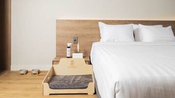 A hotel room with a wooden headboard, white bedding, a pet bed, and a small table with a spray bottle and a bowl.