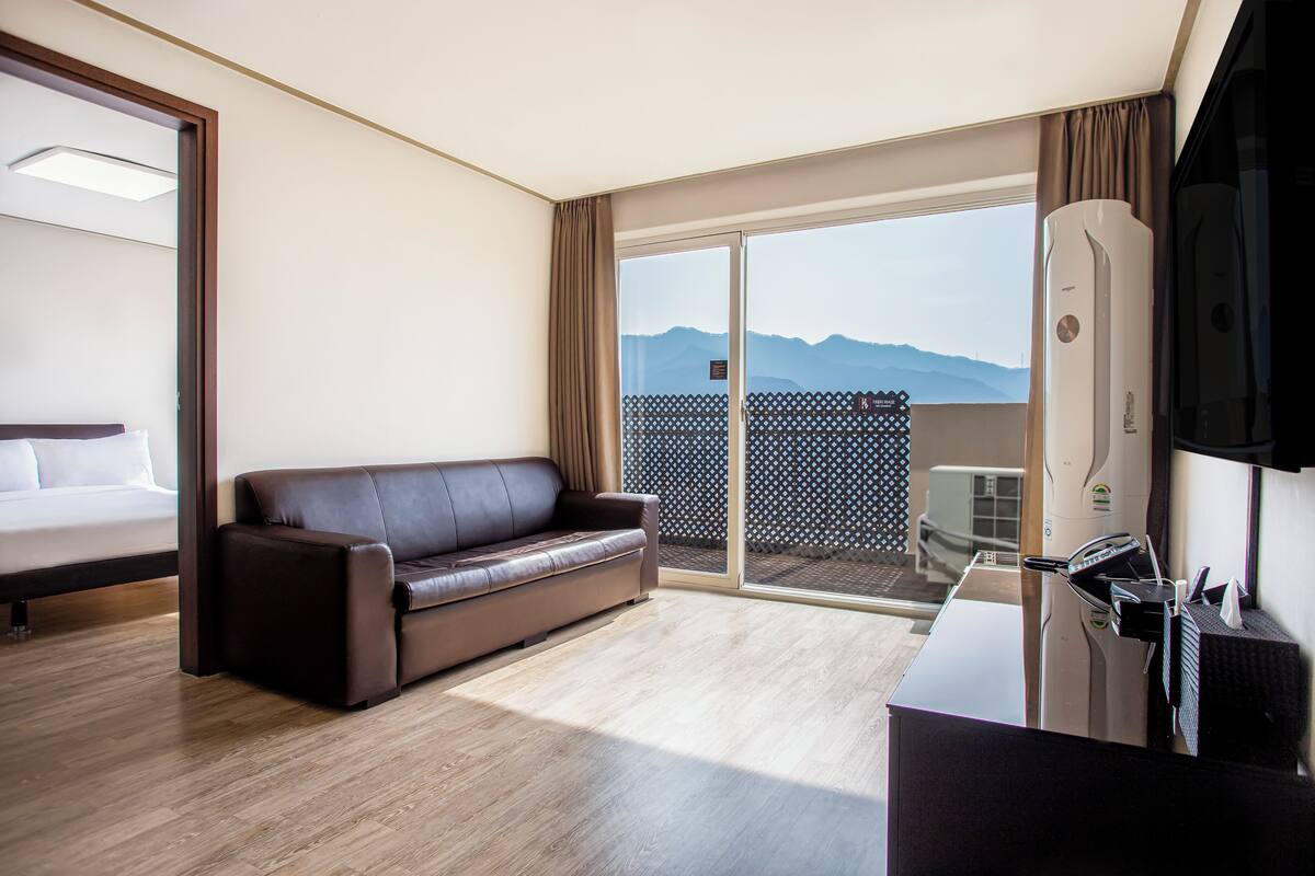 Hotelkamer met een bank, bureau, flatscreen-tv en een schuifdeur naar een balkon met uitzicht op de bergen.