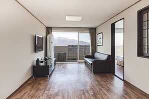 Une chambre d’hôtel moderne avec un plancher en bois, un canapé, une télévision, une petite table et une porte-fenêtre coulissante donnant sur un balcon avec vue sur la montagne.