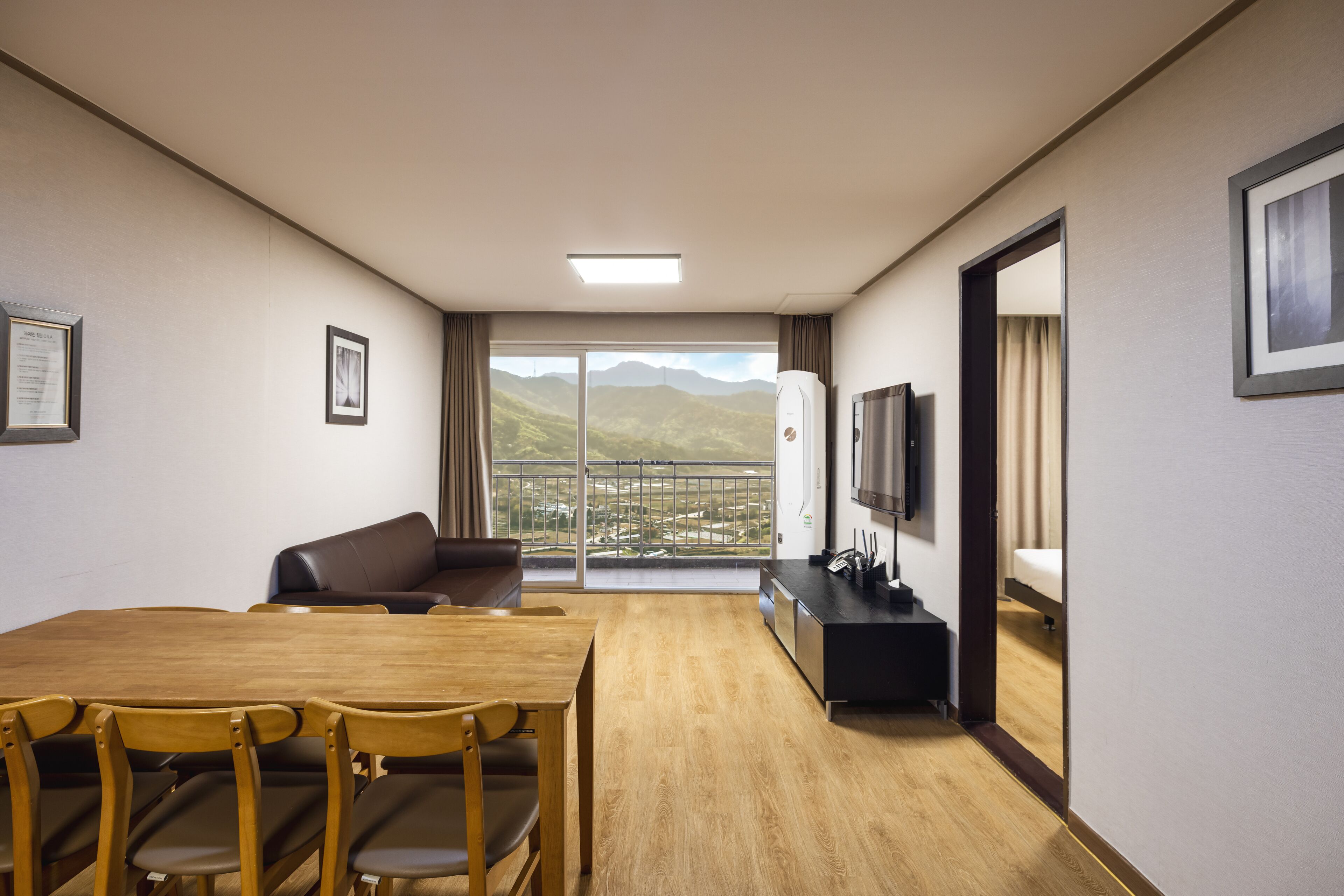 A hotel room with a wooden dining table, chairs, a sofa, a TV, and a balcony with a mountain view.