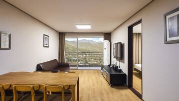 A hotel room with a wooden dining table, chairs, a sofa, a TV, and a balcony with a mountain view.