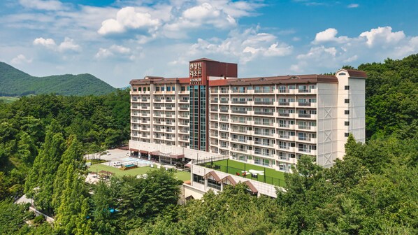 Un hôtel à plusieurs étages, doté d’une tour centrale, entouré d’une végétation luxuriante et de montagnes.