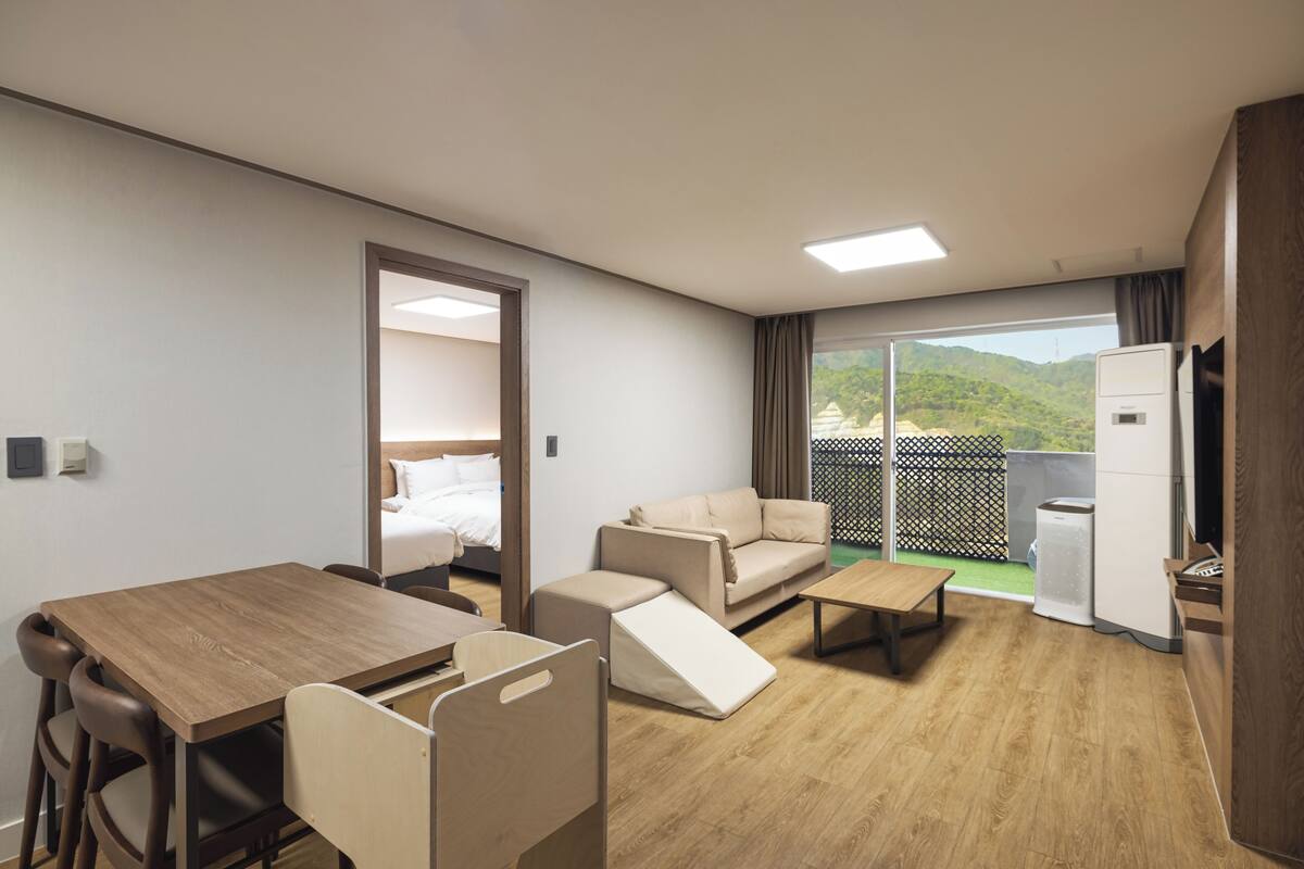 Een moderne hotelkamer met een houten eettafel, een bank, een kleinere zithoek en een bed, plus een balkon met uitzicht op groen.