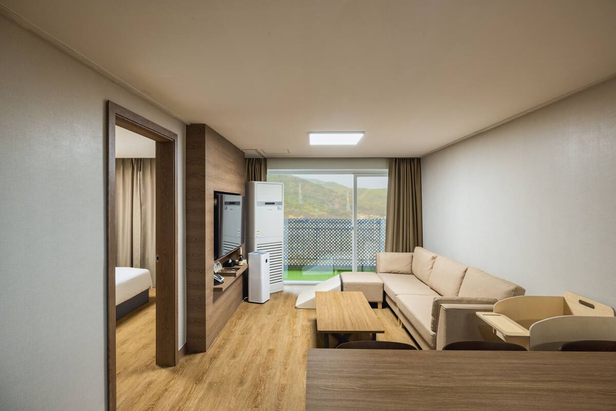 Een moderne hotelkamer met een houten vloer, een bank, een kleine tafel, een televisie en een balkon met uitzicht op groen.