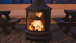 Un poêle à bois avec un feu allumé, entouré de tables de pique-nique et de gravier, le tout sur fond d’arbres et de ciel nocturne.