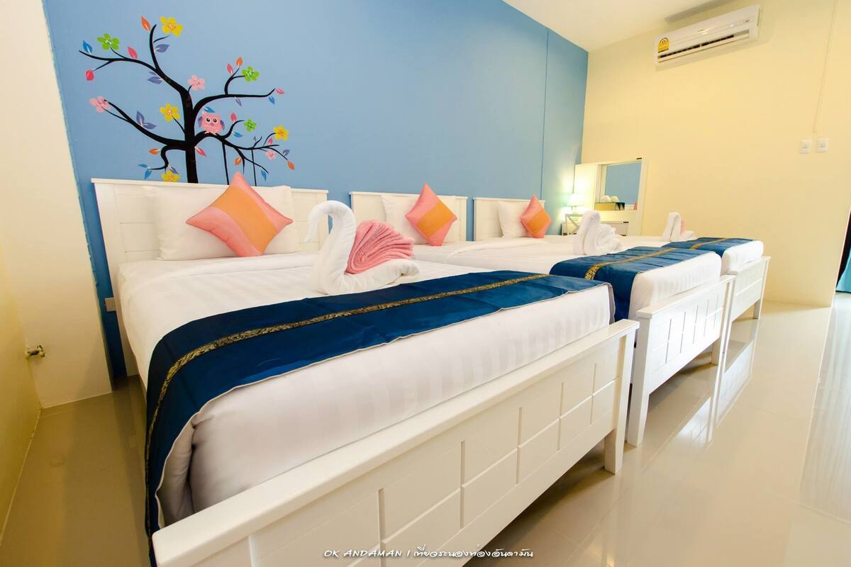 A hotel room with two beds, each with a white headboard, blue bedspreads, and pink and orange pillows. There is a wall mural of a tree with flowers and a pink swan.