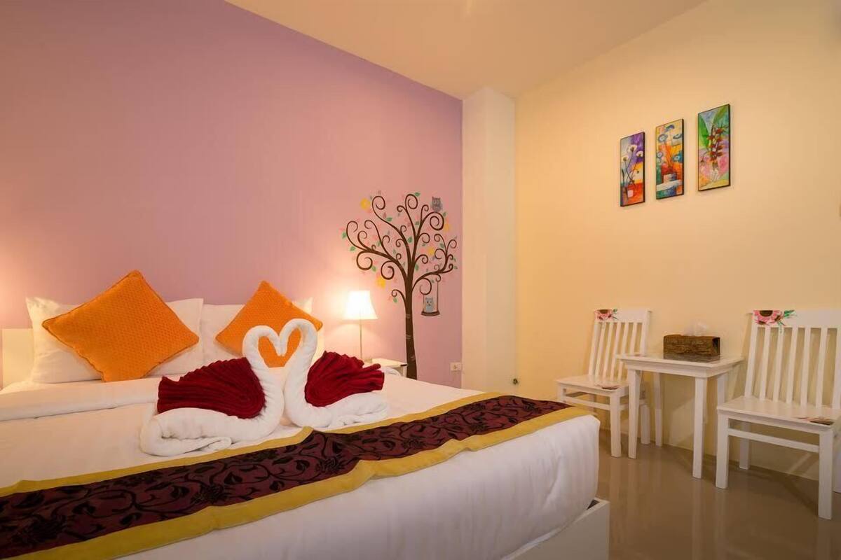 A hotel room with a bed, two white chairs, a small table, a wall-mounted tree decoration, and three framed artworks.