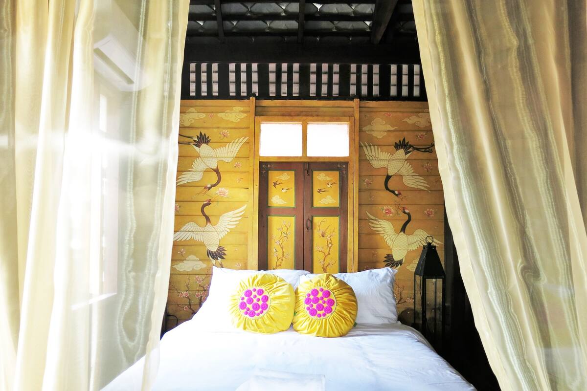 Een bed met witte beddengoed en twee kussens met roze en gele designs, voor een muur met houten panelen en vogelmotieven.