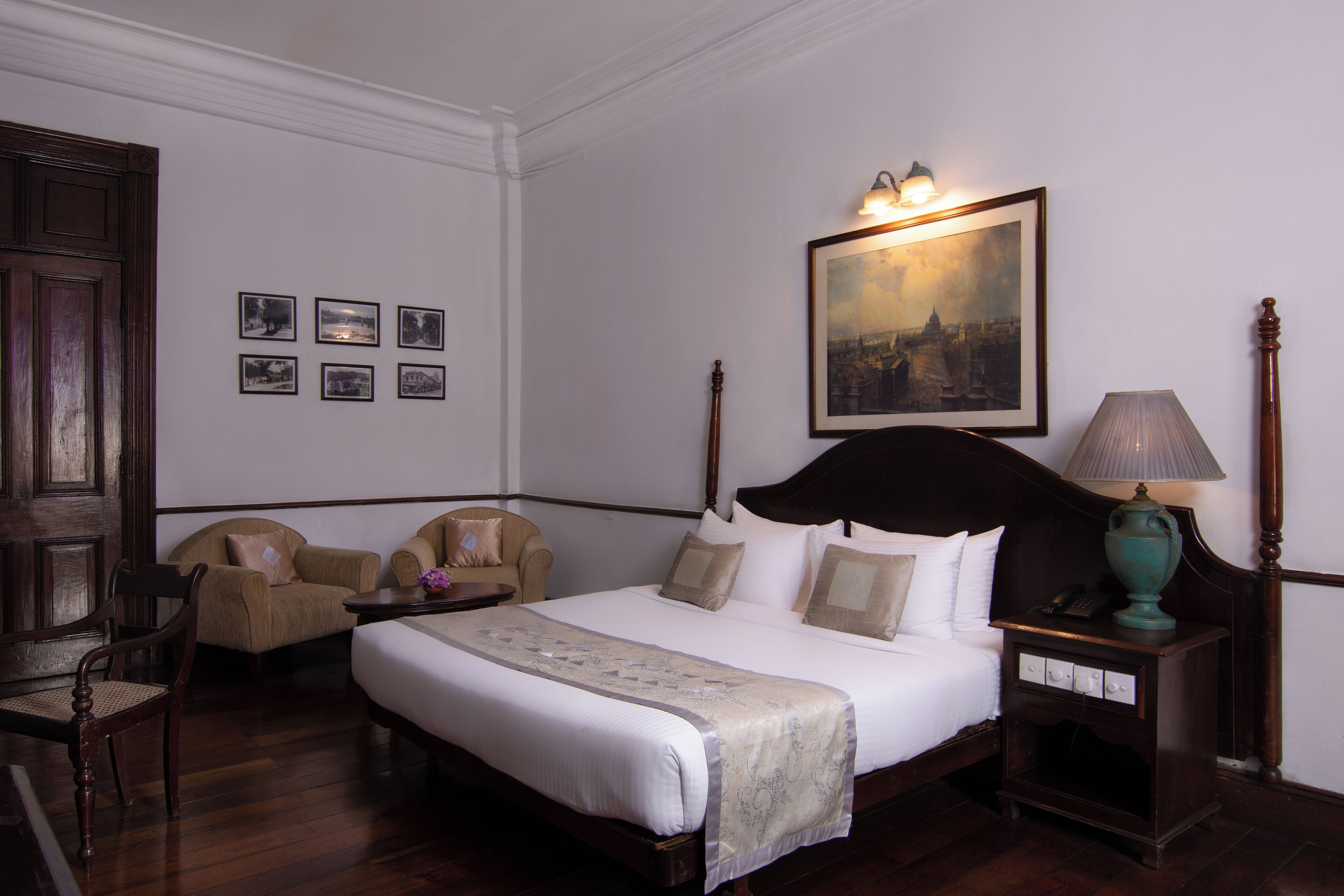 A neatly arranged bedroom with a large bed, two armchairs, a wooden nightstand with a lamp, and a framed cityscape painting on the wall.