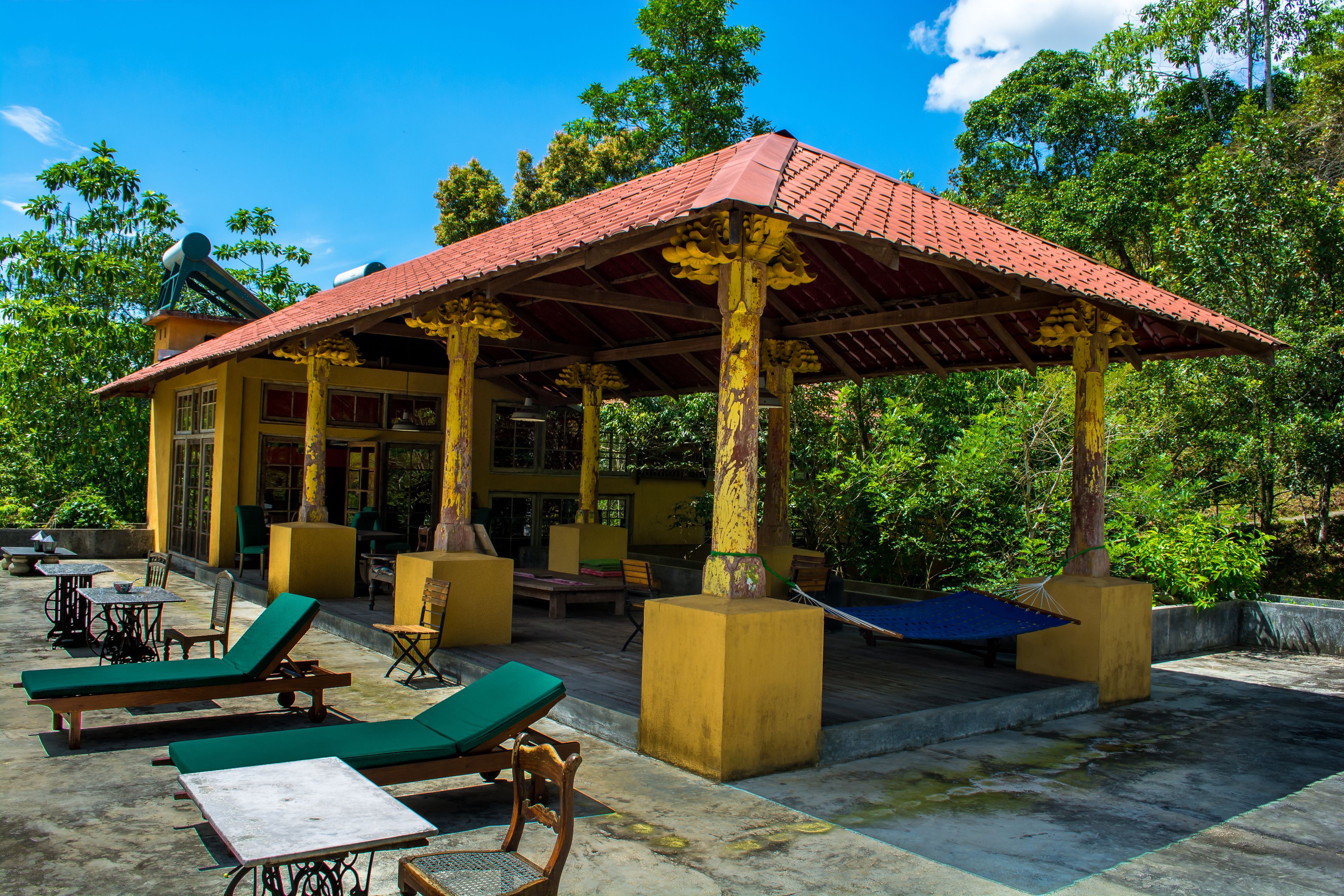 A covered outdoor seating area with lounge chairs and tables, surrounded by greenery.