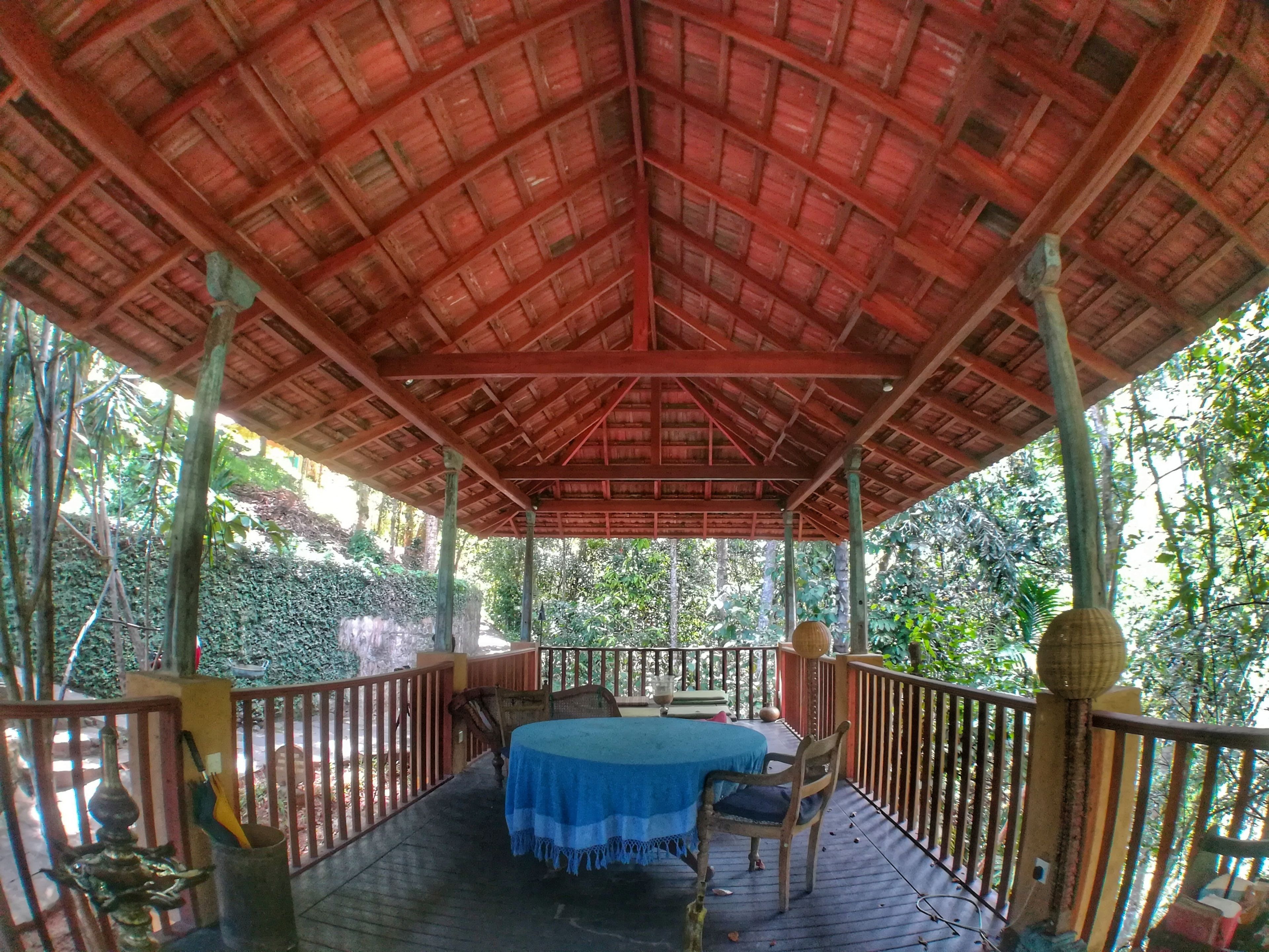 Un espace extérieur abrité, aménagé avec une table et des chaises, entouré d’une terrasse en bois et de rampes, avec des arbres en arrière-plan.